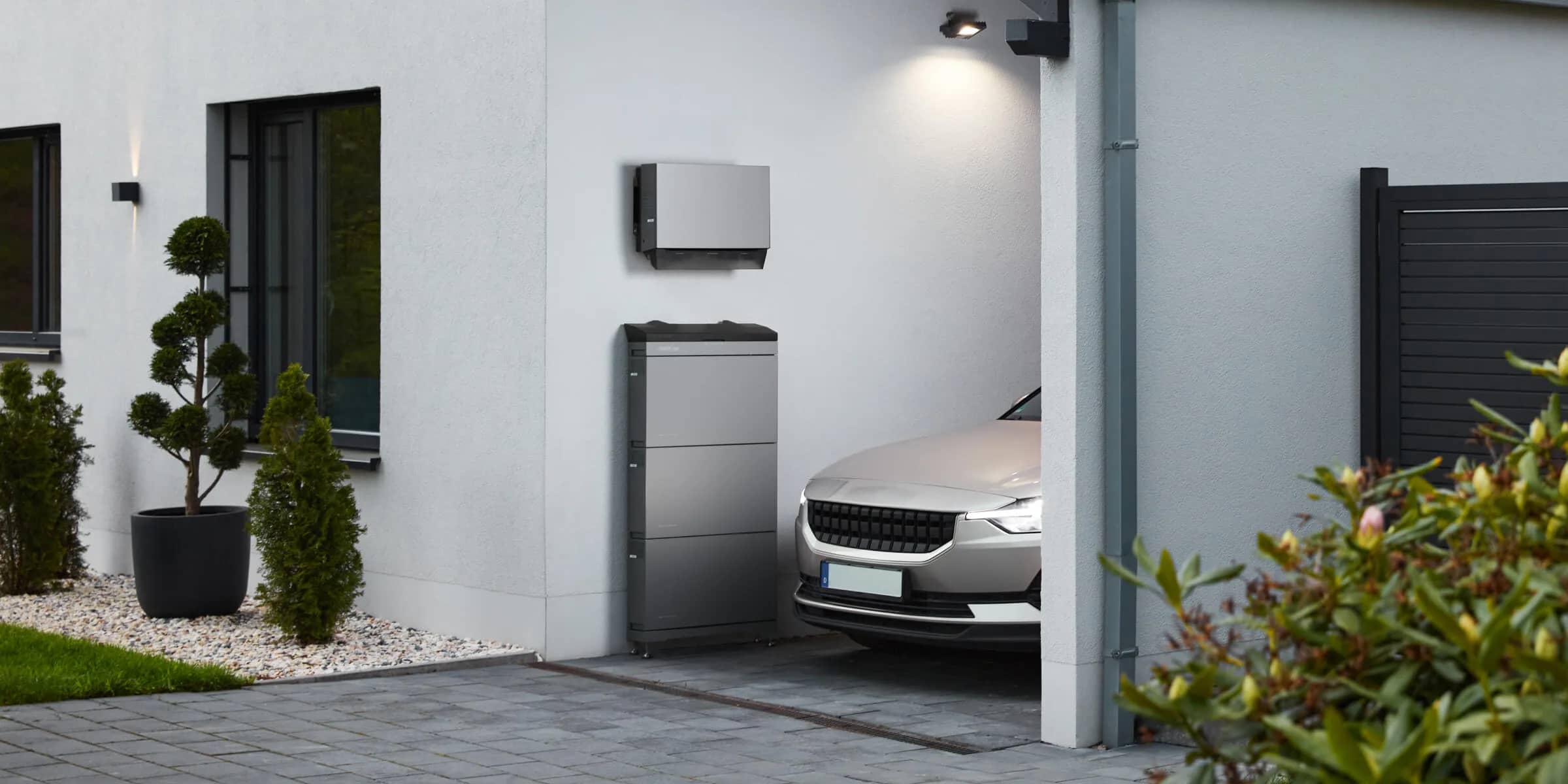 A home battery and inverter on the white wall of a house beside the drive, with a car parked alongside